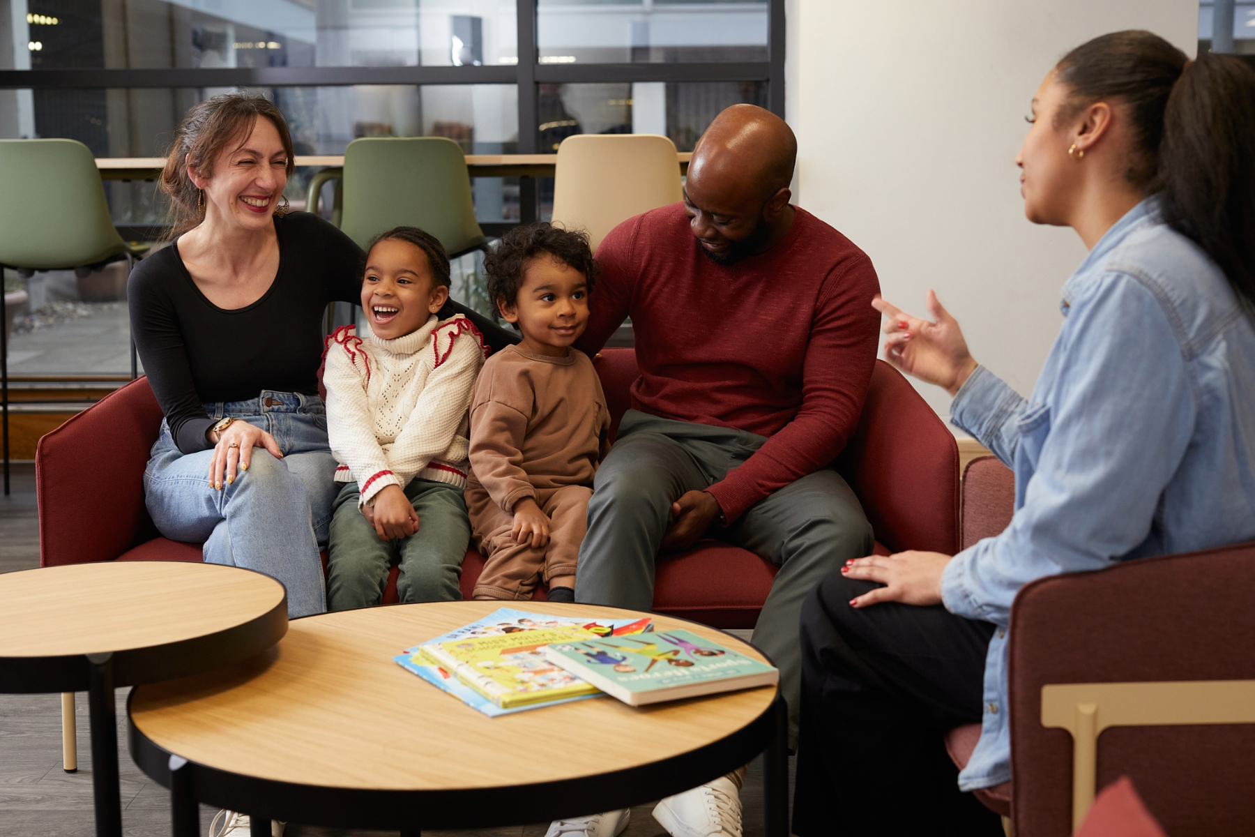 A mixed heritage family sit on a sofa laughing within a Local Authority lounge area with a female social worker.