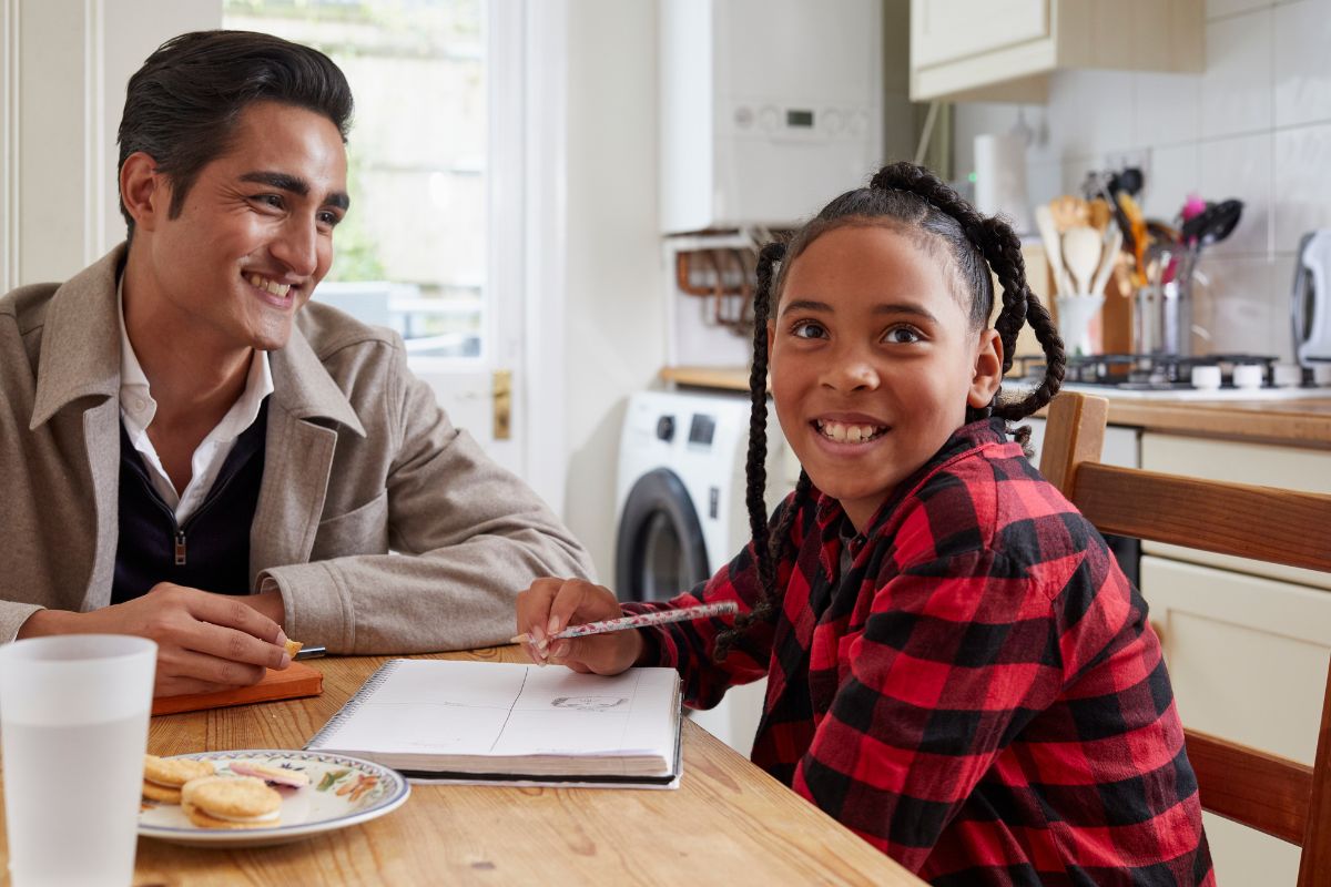 A male social worker sits at a kitchen table with a young girl, smiling